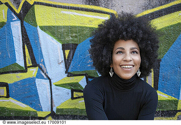 Fröhliche junge Frau mit Afrolook  die an einer Graffitiwand sitzt