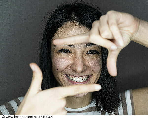 Fröhliche junge Frau schaut durch den Finger vor grauem Hintergrund