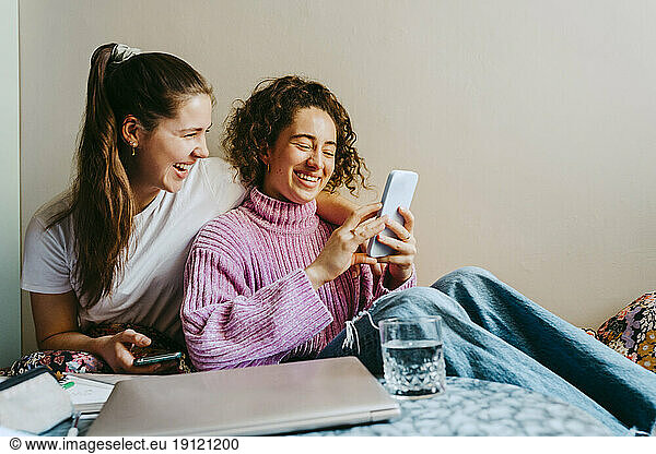 Fröhliche junge Freundinnen teilen sich ein Smartphone  während sie zu Hause sitzen