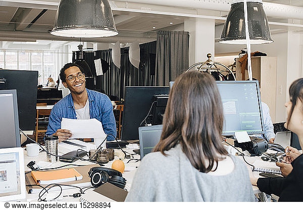 Fröhliche multiethnische IT-Fachleute unterhalten sich am Schreibtisch in einem kreativen Büro
