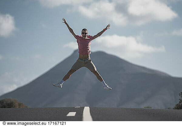 Fröhlicher junger männlicher Tourist  der an einem sonnigen Tag auf Lanzarote über die Straße gegen vulkanisches Gestein springt  Spanien