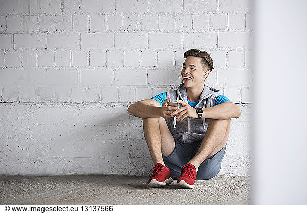 Fröhlicher männlicher Athlet hält Smartphone in der Hand  während er auf dem Gehweg sitzt
