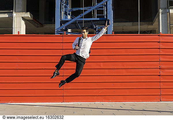 Fröhlicher männlicher Unternehmer mit erhobenem Arm  der auf dem Fußweg gegen ein Bauwerk springt