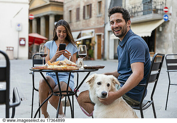 Fröhlicher Mann sitzt mit Hund und Frau in einem Straßencafé