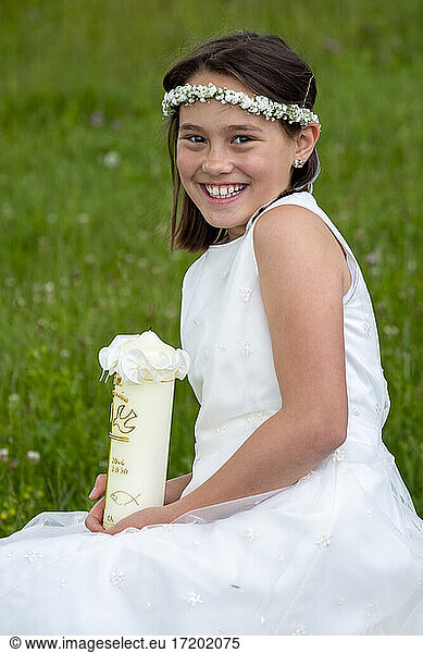Fröhliches Mädchen in weißem Kleid hält Kerze am Tag der Kommunion