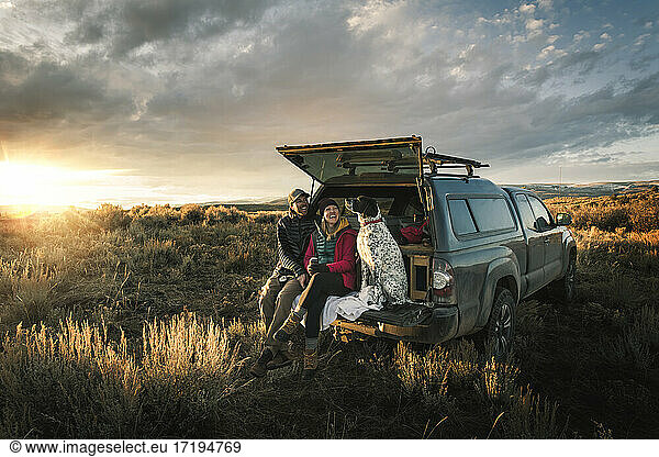 Fröhliches Paar mit Hund im Kofferraum eines Geländewagens auf einem Feld im Urlaub