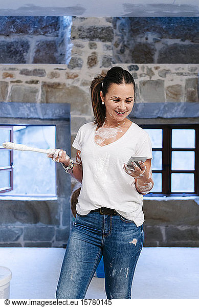 Frau benutzt ihr Handy  während sie die Wände ihres neuen Hauses streicht