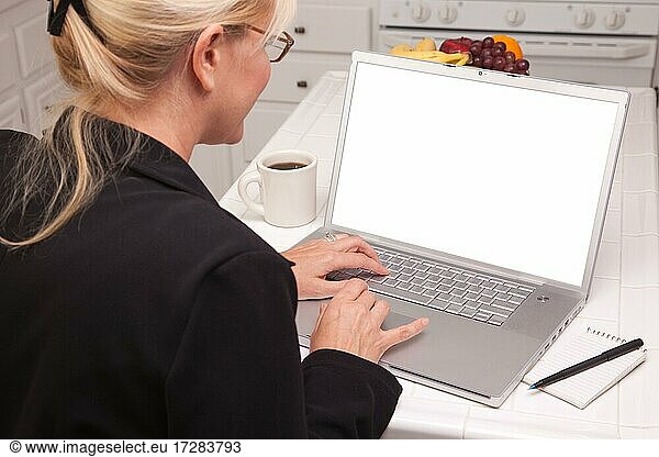 Frau  die in der Küche sitzt und einen Laptop mit leerem Bildschirm benutzt. Der Bildschirm kann mit dem mitgelieferten Beschneidungspfad leicht für eine eigene Nachricht oder ein Bild verwendet werden