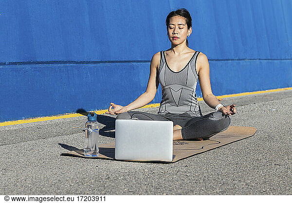 Frau im Lotussitz lernt Yoga  während sie vor einem Laptop sitzt