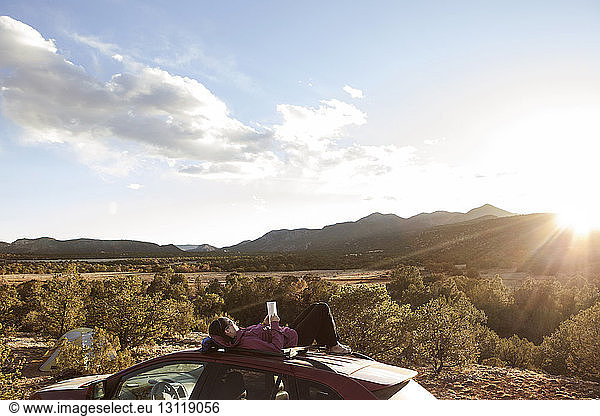 Frau liest Buch  während sie bei Sonnenschein auf der Motorhaube eines Autos gegen einen Berg liegt