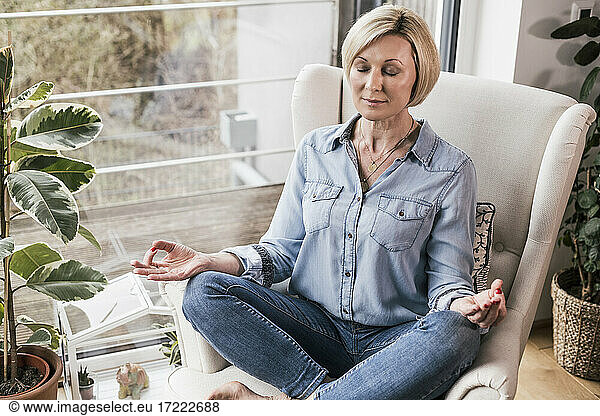 Frau mit geschlossenen Augen sitzt im Lotussitz auf einem Sessel zu Hause