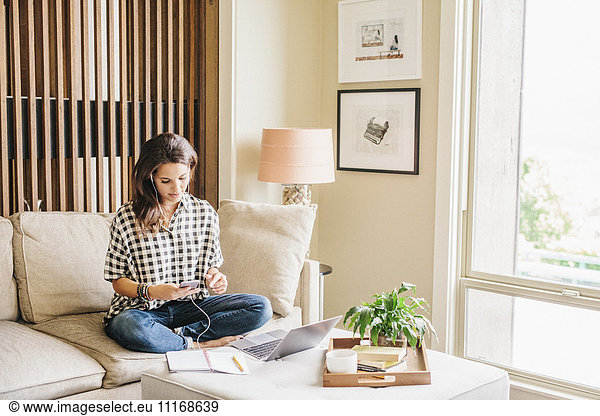Frau mit langen braunen Haaren sitzt auf einem Sofa mit Laptop und Notebook und arbeitet.