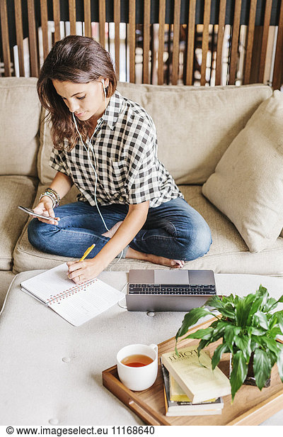 Frau mit langen braunen Haaren sitzt auf einem Sofa mit Laptop und Notebook und arbeitet.