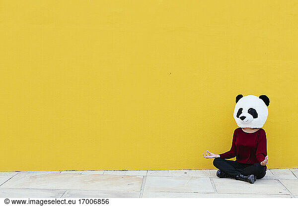 Frau mit Panda-Maske sitzt im Lotussitz an einer gelben Wand