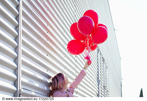 Frau mit rosa Haaren hält einen Strauß rosa Luftballons vor einer Metallwand