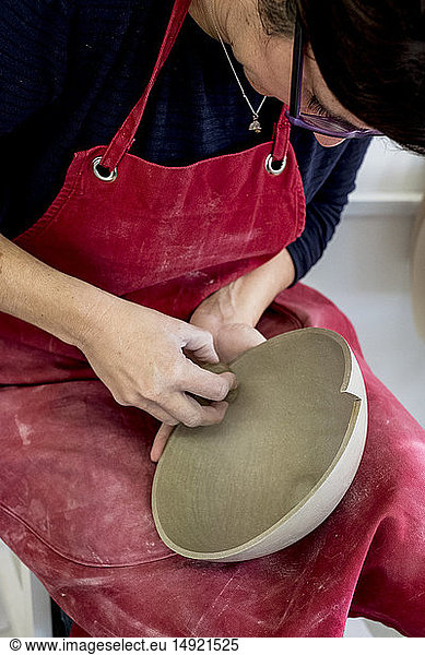 Frau mit roter Schürze sitzt in ihrer Keramikwerkstatt und arbeitet an einer Tonschüssel.