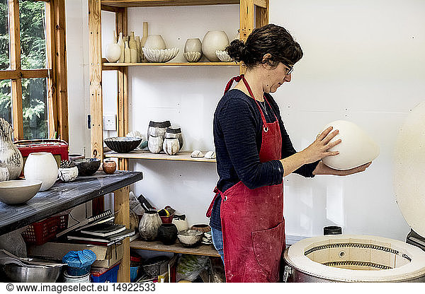 Frau mit roter Schürze steht in ihrer Werkstatt neben dem Brennofen und hält eine Keramikvase.