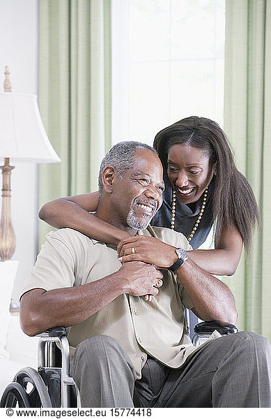 Frau mittleren Alters umarmt einen Mann mittleren Alters von hinten  der im Rollstuhl sitzt und lächelt