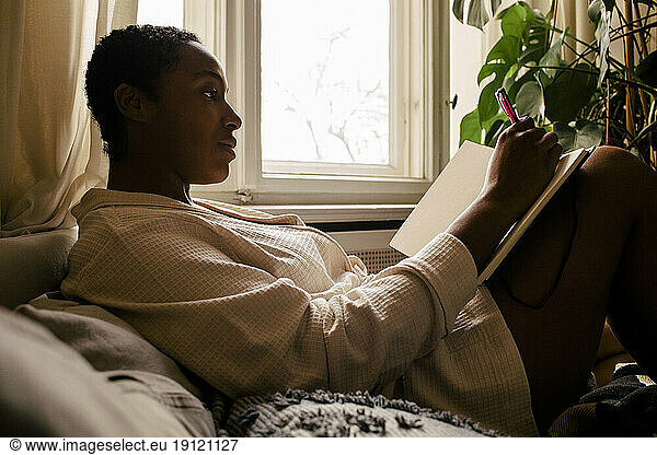 Frau schreibt in einem Buch  während sie sich zu Hause auf dem Bett ausruht
