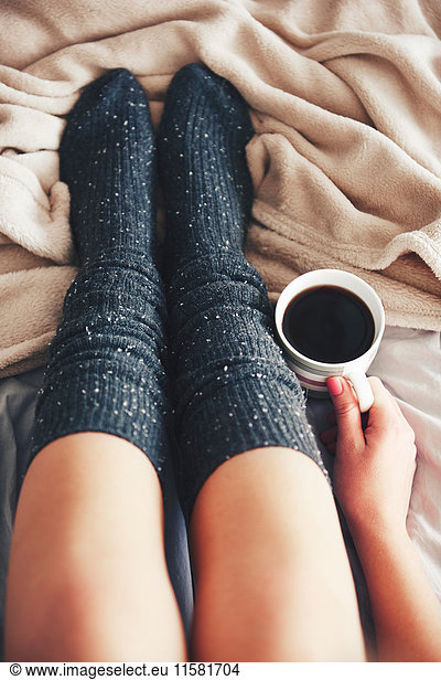 Frau sitzt auf dem Bett  trägt warme Socken  hält eine Tasse Kaffee  niedriger Schnitt  Draufsicht