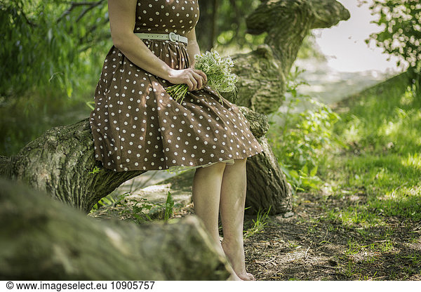 Frau sitzt auf einem Baumstamm und hält einen Strauß gepflückter Blumen  Teilansicht
