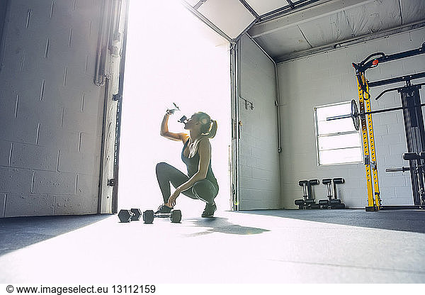 Frau trinkt Wasser  während sie in hell erleuchteter Turnhalle kauert