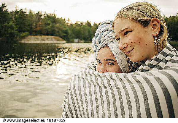 Frau umarmt Freundin in Handtuch gewickelt am See im Urlaub