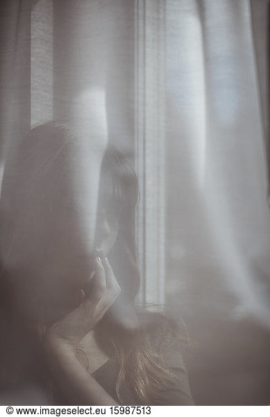 Frau versteckt sich hinter weißem Fenstervorhang