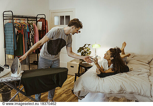 Frau zeigt dem Mann  der zu Hause im Schlafzimmer Wäsche bügelt  ihr Smartphone