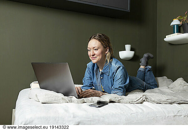 Freelancer in denim shirt working with laptop lying on bed at home