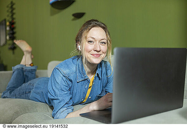 Freelancer in denim shirt working with laptop lying on couch