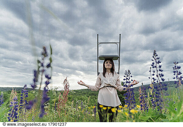 Freelancer meditating with chair on head in meadow