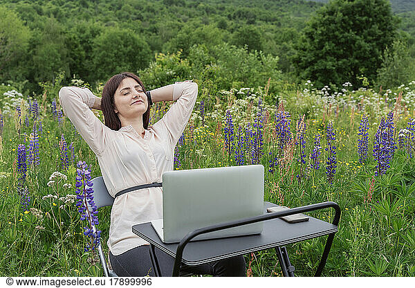 Freelancer relaxing with hands behind head at desk in lupine meadow
