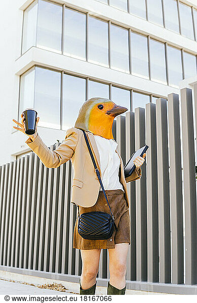 Freelancer wearing bird mask holding coffee cup and tablet PC by office building