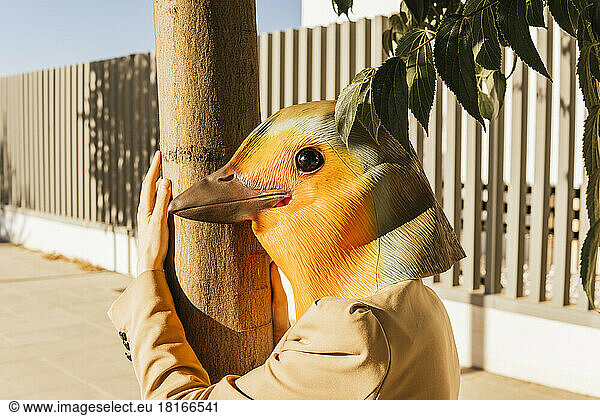 Freelancer wearing bird mask hugging tree at footpath