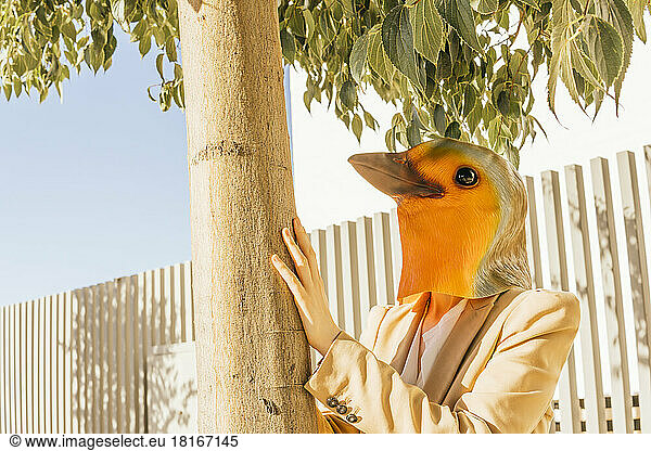 Freelancer wearing bird mask standing by tree on sunny day