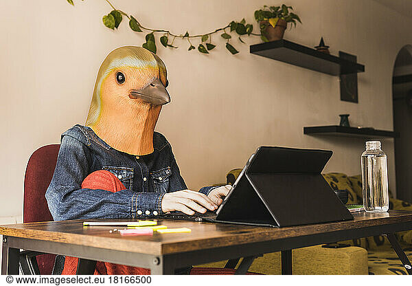 Freelancer wearing bird mask using tablet PC sitting at home