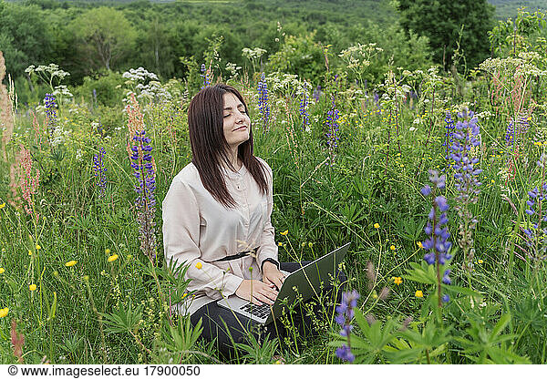Freelancer with laptop and eyes closed sitting in meadow