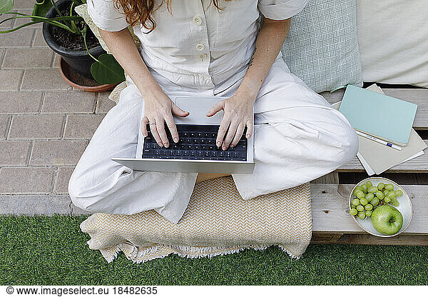 Freelancer working on laptop sitting cross-legged on crate at backyard