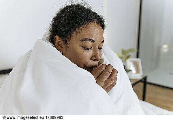 Freezing young woman sitting on the bed wrapped in a blanket rubbing hands