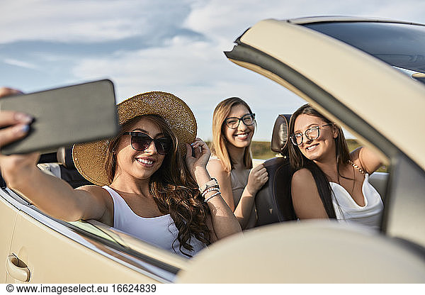 Freunde  die ein Selfie auf einem Smartphone machen  während sie in einem Luxusauto sitzend einen Road Trip genießen