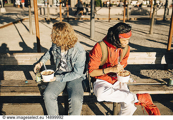Freunde essen Essen auswärts,  während sie an sonnigen Tagen in der Stadt auf einer Bank sitzen