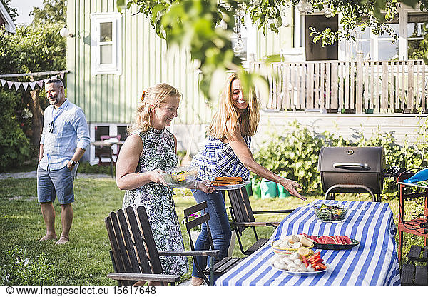 Freundinnen arrangieren Essen auf dem Tisch für geselliges Beisammensein im Garten am Sommerwochenende