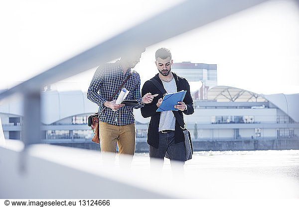 Friends discussing over document while walking against clear sky