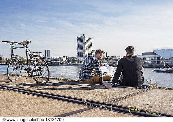 Friends looking at document while sitting by bicycle on promenade against clear sky