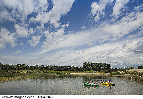Friends relaxing on pool rafts on lake against cloudy sky
