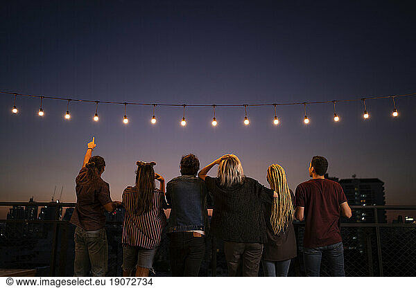 Friends spending leisure time under string lights on rooftop at dusk