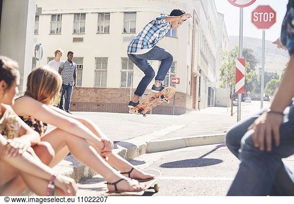Friends watching teenage boy jumping skateboard at sunny urban corner