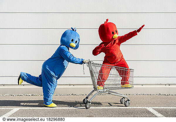 Friends wearing red and blue duck costumes having fun with shopping cart by wall