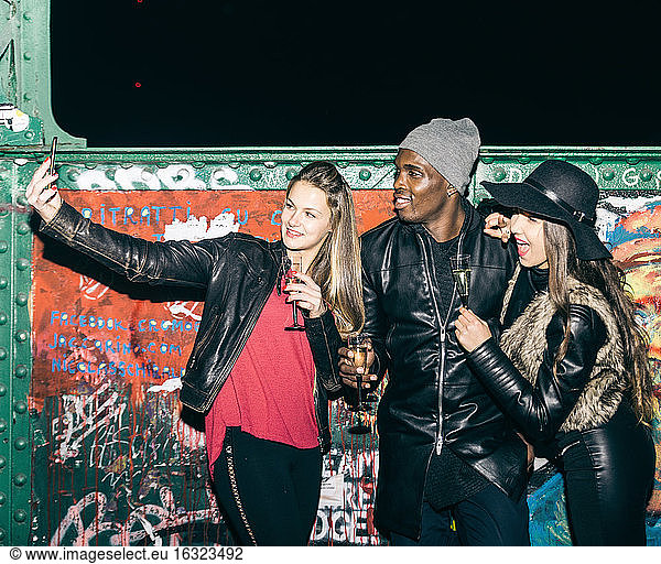 Friends with champagne glasses taking a selfie at graffiti wall at night